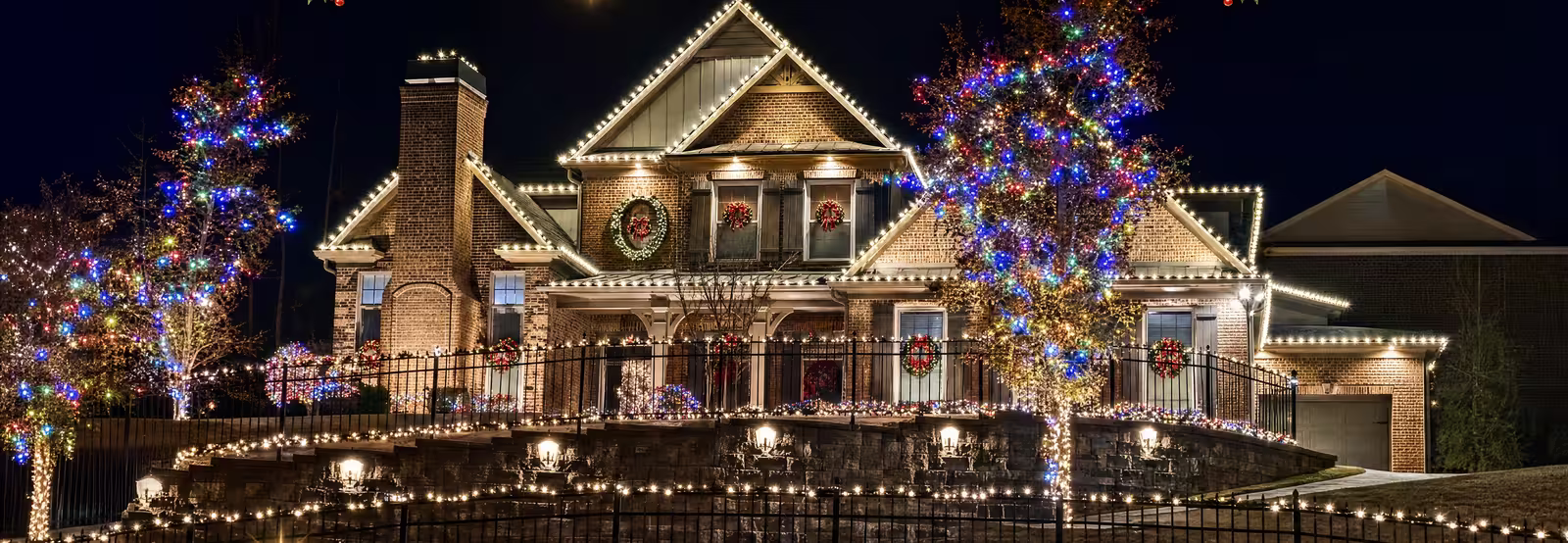 A two-story brick house at night with every gable and roofline outlined in warm-white bulbs, trees wrapped in colored lights, and a lit stone retaining wall along the front walk.